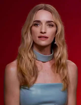 Head and shoulders photograph of a woman with long wavy blonde hair and a pale blue high-necked top against a red background.