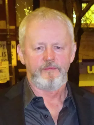 A grey-haired man with a short grey beard, in a dark open-collar shirt and jacket, photographed outdoors at night.