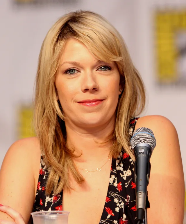 Mary Elizabeth Ellis with shoulder-length blonde hair, in a floral top, seated at a panel microphone with a convention backdrop behind her.