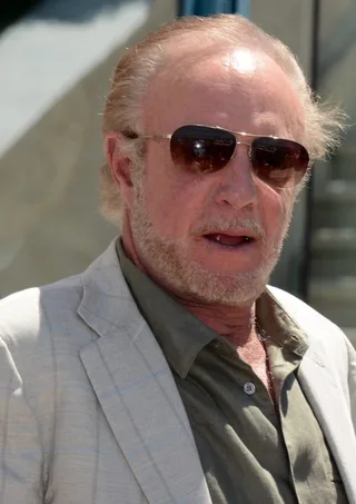Close photograph of an older man with a short grey beard and tinted aviator sunglasses, wearing a pale jacket over an olive shirt, outdoors in sunlight.