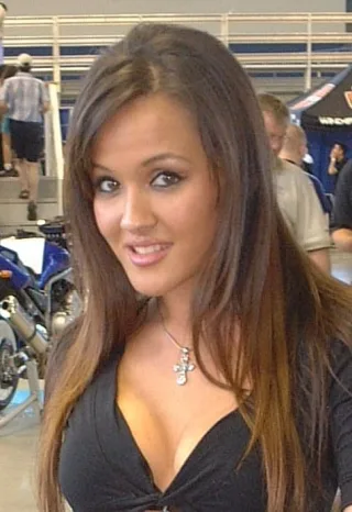 Close photograph of a smiling woman with long brown hair and a cross pendant, wearing black, at an indoor motorsport event.