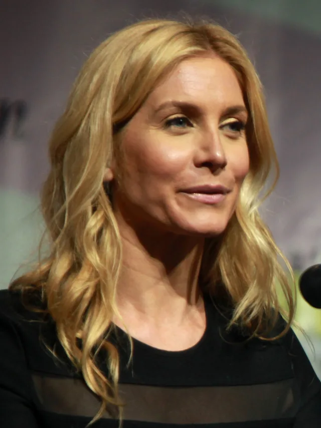 Close portrait of a woman with long wavy blonde hair and a black top, smiling and looking to one side in front of a blurred backdrop.