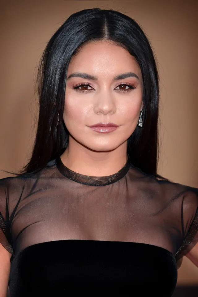 Head and shoulders photograph of a woman with long straight dark hair, wearing a black top with sheer sleeves and a drop earring, against a warm tan backdrop.