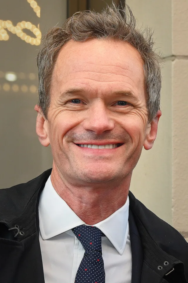 Close photograph of a smiling man with short tousled grey-brown hair, wearing a dark jacket and a white shirt with a blue polka-dot tie.
