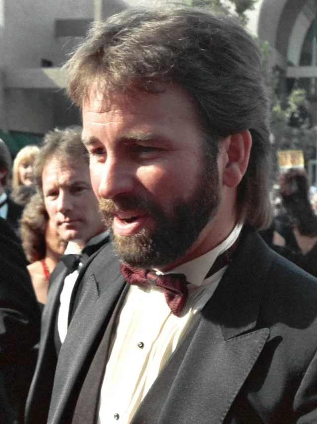 A bearded man with shoulder-length brown hair in a black dinner jacket and burgundy bow tie, photographed mid-sentence outdoors at an awards event.
