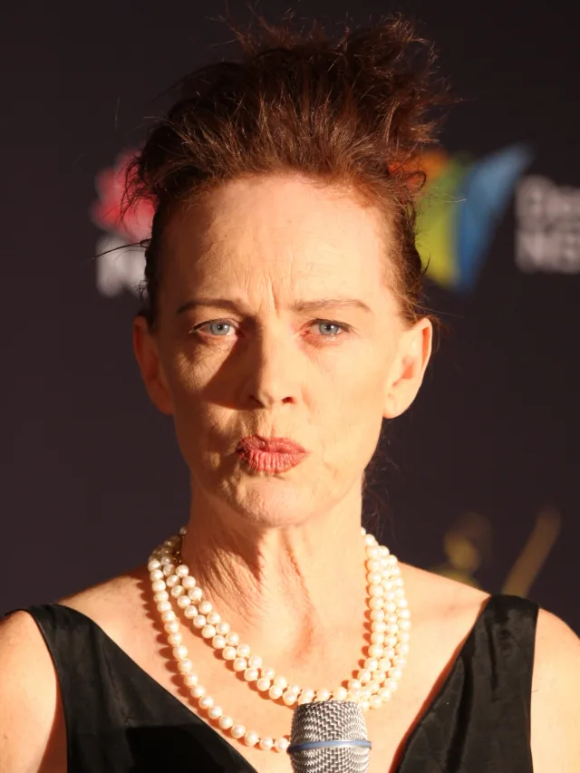 Close portrait of Judy Davis at a microphone, wearing a black top and a triple strand of pearls, with swept-up auburn hair.