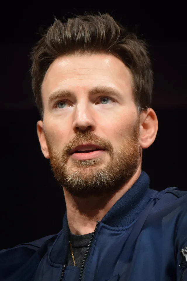 A bearded man with short brown hair in a navy zip-up jacket, looking attentively to the side against a dark background at a convention panel.