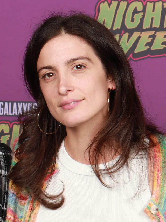 A woman with long dark brown hair and gold hoop earrings, in a white top and a colourful knitted cardigan, looking at the camera with a slight smile.