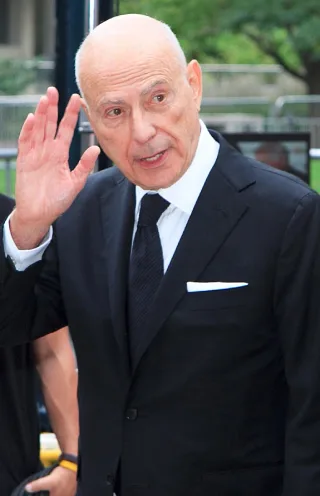 Alan Arkin, a bald older man in a dark suit and tie with a white pocket square, raising one hand in greeting outdoors.