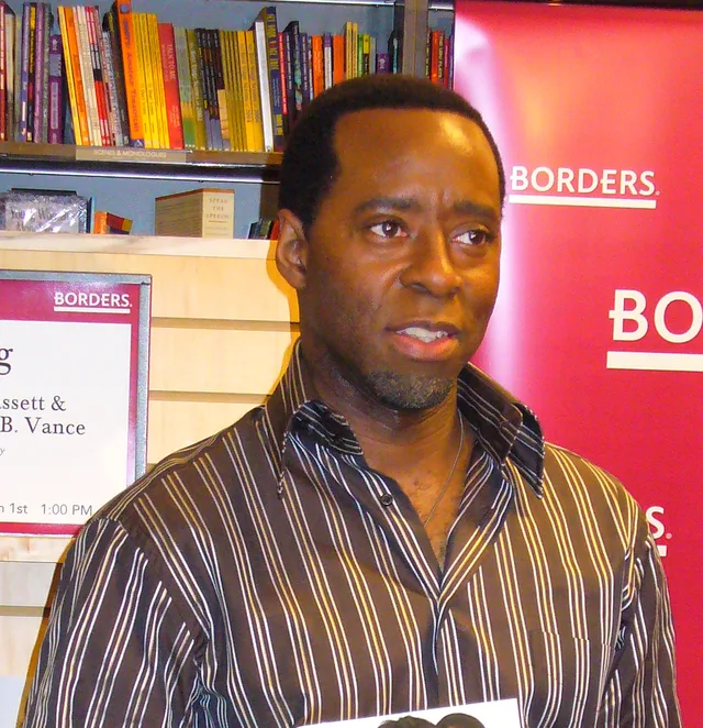 A man with short dark hair and a grey goatee in a brown striped shirt, looking to the side in front of bookshelves at a bookshop event.