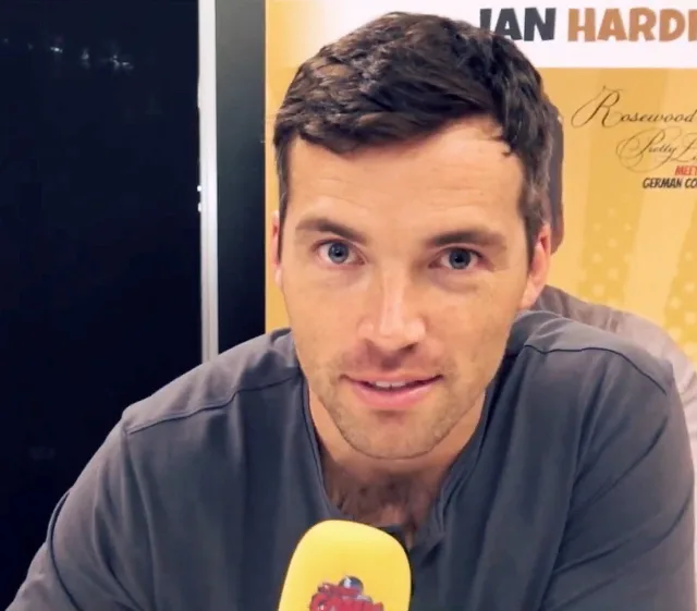 Close-up photograph of a smiling man with short dark hair and light eyes, wearing a grey top and speaking into a yellow microphone at a fan convention.
