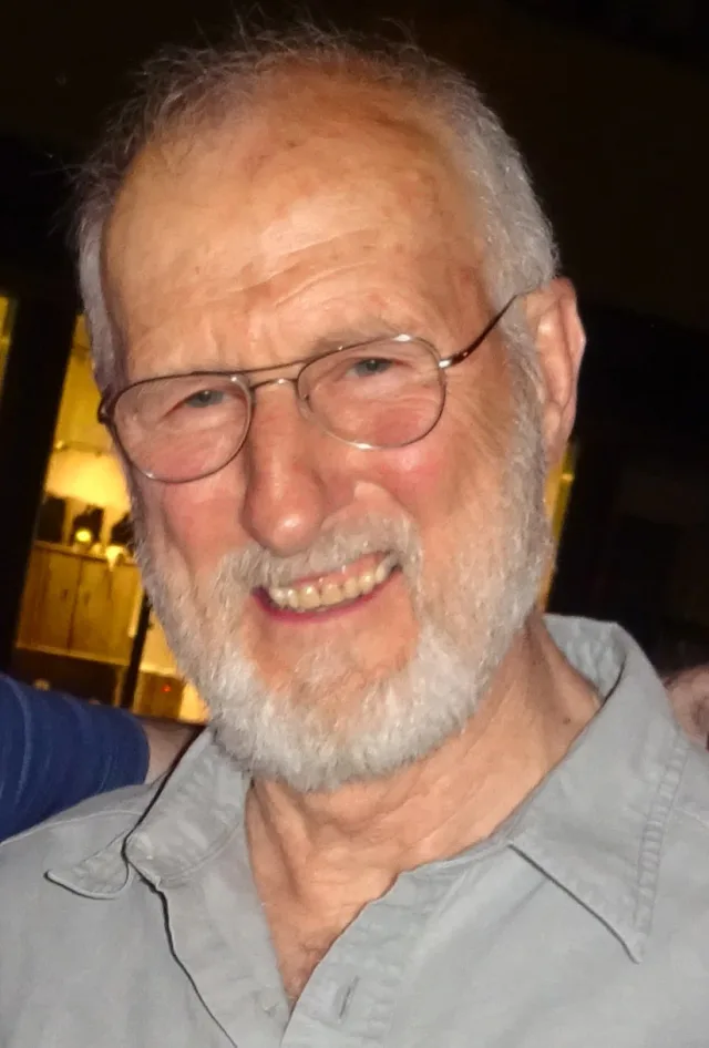 Close portrait of James Cromwell, an older man with grey hair and a white beard, wearing round glasses and a grey collared shirt, smiling.