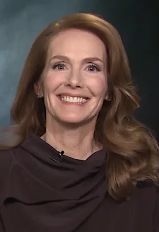 Smiling woman with long auburn hair and a dark brown draped top, shown in a head-and-shoulders interview frame.