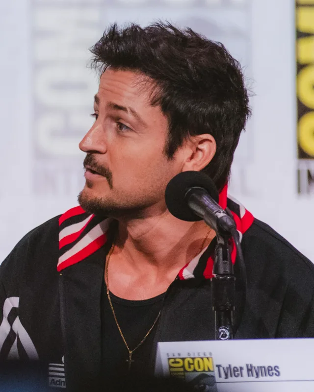 Tyler Hynes in profile at a convention panel table, dark hair and short beard, speaking into a microphone.
