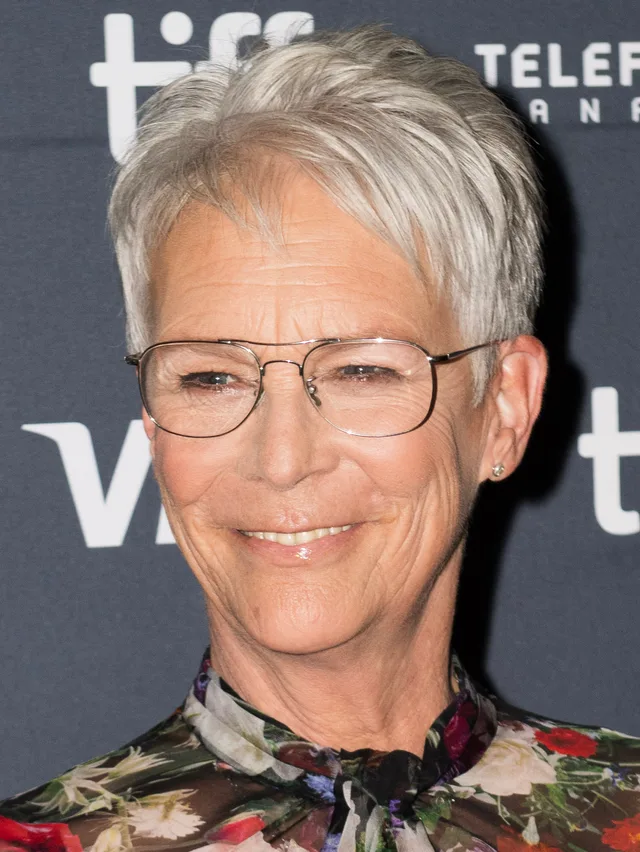 Close colour photograph of a smiling woman with short silver hair and thin metal-framed glasses, wearing a floral blouse, in front of a dark festival backdrop.