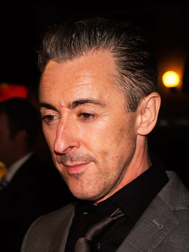 Close portrait of Alan Cumming looking down, with slicked-back dark hair, a black shirt and tie and a grey jacket, in low light.