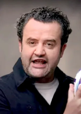 Close photograph of Daniel Mays with dark curly hair and stubble, wearing a dark shirt and holding a ball while speaking.