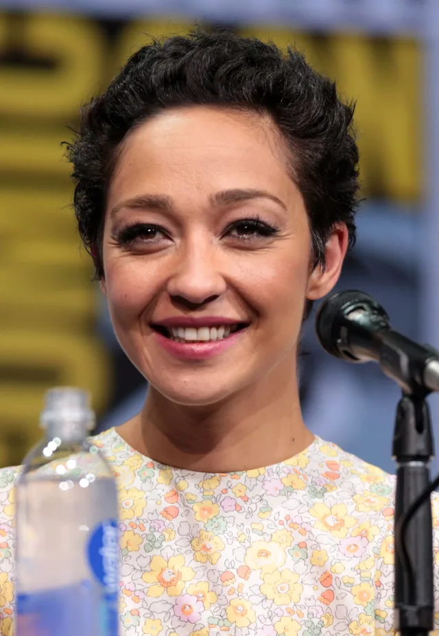 Smiling woman with short dark curly hair in a floral top, seated at a panel table with a microphone in front of her.