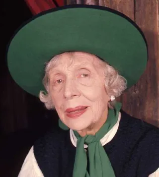 Portrait of an elderly woman with curled white hair in a wide green hat and a green scarf over a dark waistcoat and white blouse.