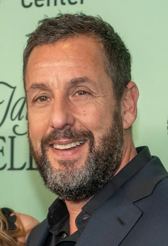 Close-up photograph of a smiling man with short dark hair and a grey-flecked beard, wearing a dark jacket, against a pale green backdrop.