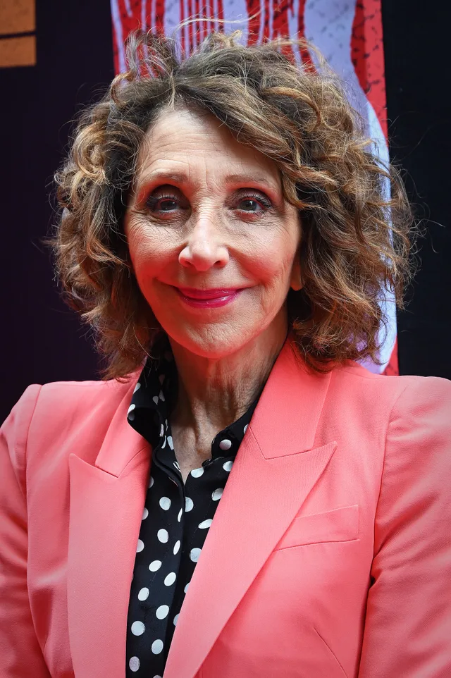 Head and shoulders of a smiling woman with wavy curly brown hair, wearing a coral blazer over a black polka dot blouse.