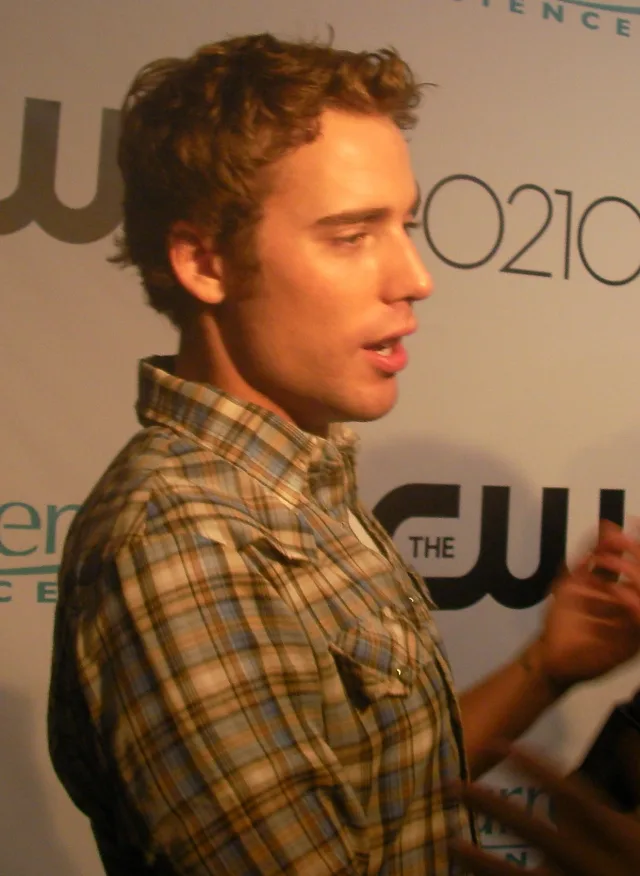 Dustin Milligan in profile, speaking, with tousled light brown hair and a plaid shirt, in front of a press backdrop.