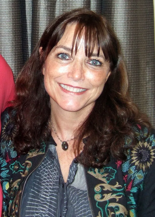 Smiling woman with long dark brown hair and a fringe, wearing a pendant necklace and an embroidered dark jacket, in front of a curtain.