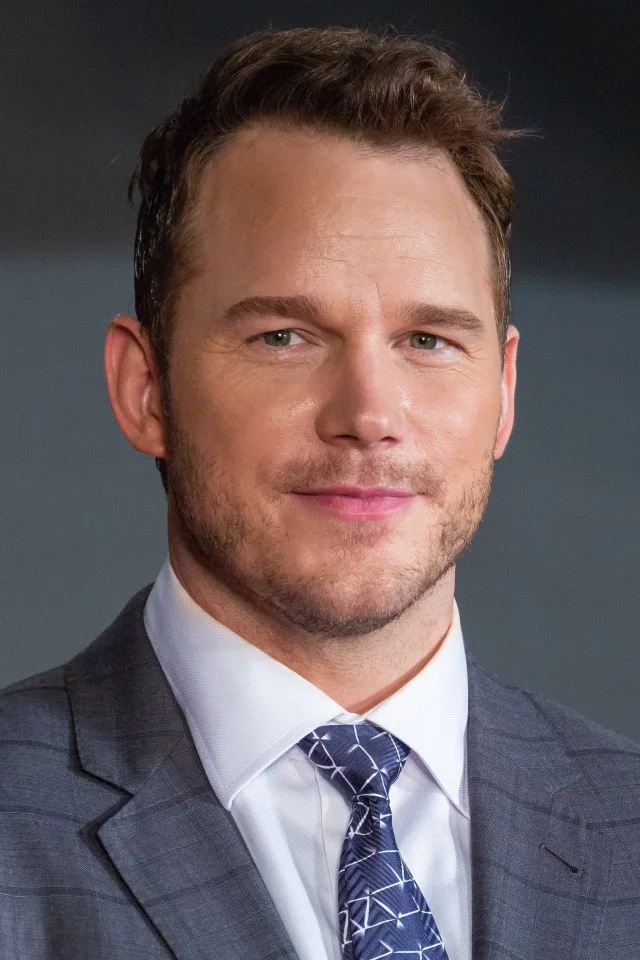 Close portrait of a smiling man with short brown hair and light stubble, wearing a grey checked suit jacket, white shirt and blue patterned tie.