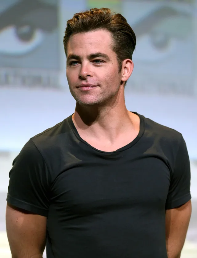 A man with short swept-back brown hair and light stubble, in a dark grey t-shirt, looking off to the side with a slight smile in front of a pale blurred backdrop.