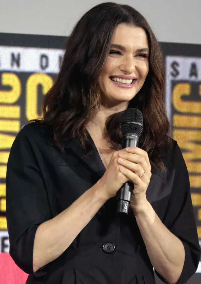 Laughing woman with long dark wavy hair in a black collared top, holding a microphone in both hands on a convention stage.