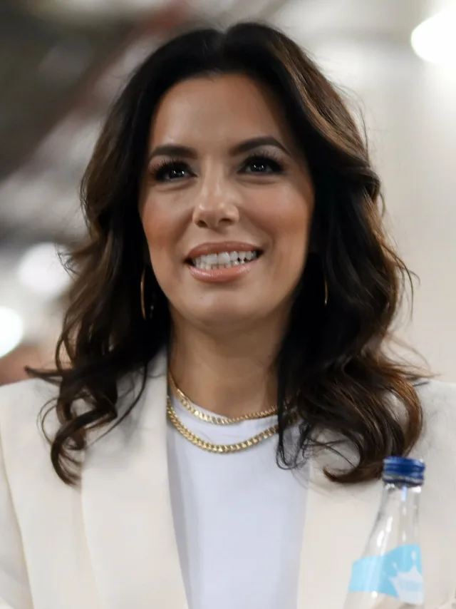 Smiling woman with long wavy dark hair, wearing a cream blazer, white top and layered gold chain necklaces, with hoop earrings.