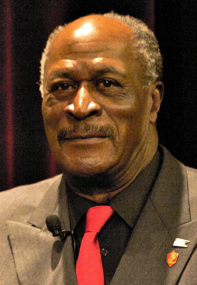 Close portrait of an older man with grey hair and a moustache, wearing a grey jacket, dark shirt and red tie with a small clip microphone.