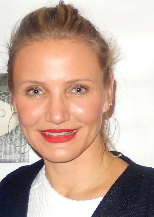 Close portrait of Cameron Diaz smiling, with red lipstick, hair tied up, in a navy cardigan over a white top.