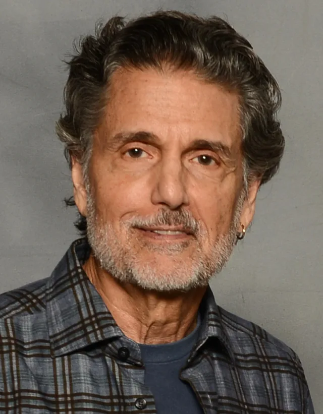 Man with dark wavy hair and a grey beard, wearing a blue plaid shirt and a small gold earring, smiling slightly at the camera.