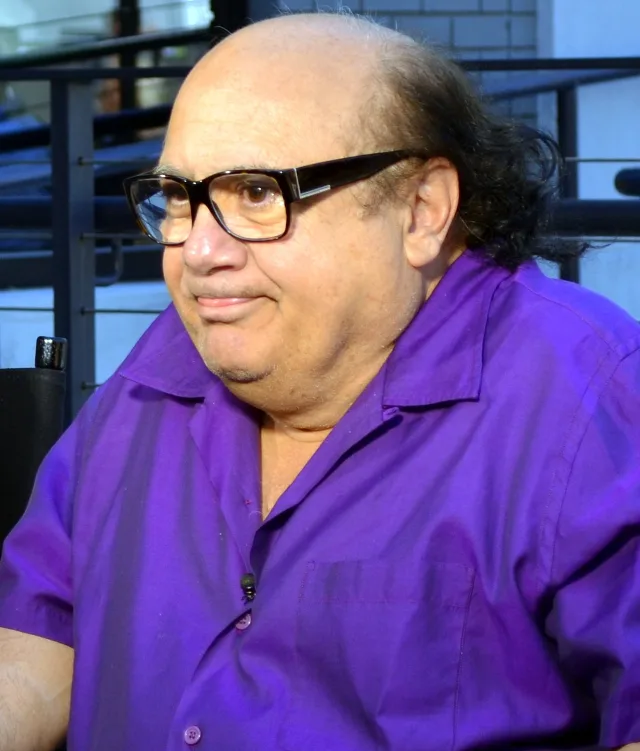 Danny DeVito in a purple shirt and dark-framed glasses, seated outdoors and looking to one side.