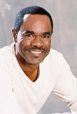 Colour headshot of a smiling man with short dark hair and a moustache, wearing a cream ribbed top against a grey backdrop