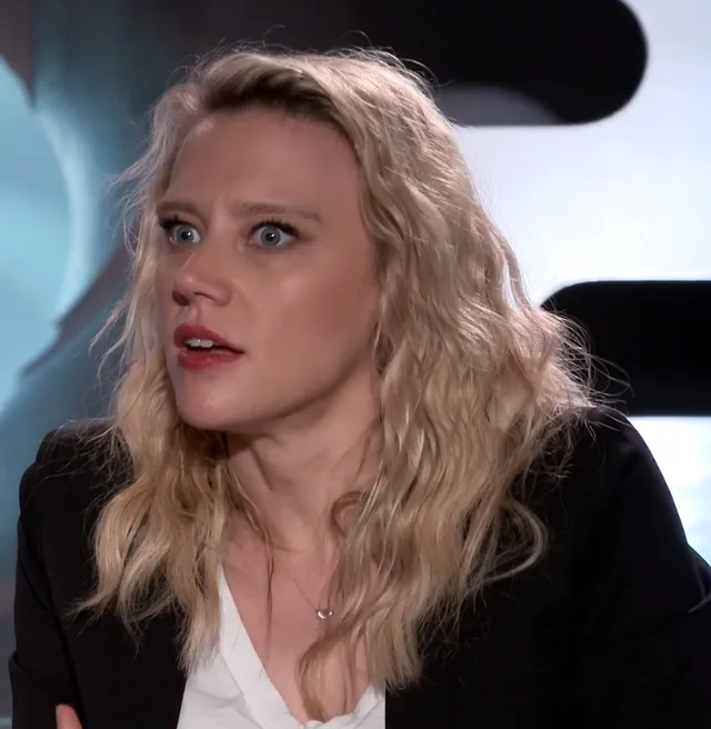 A woman with wavy blonde hair and red lipstick, in a black blazer over a white top, glancing to one side during an interview.