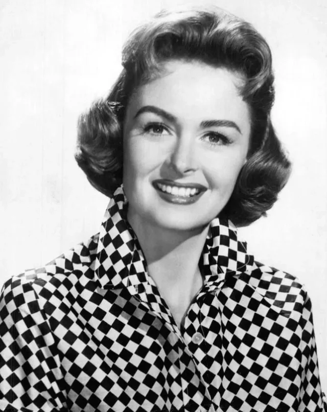 Black and white studio portrait of a smiling woman with wavy hair, wearing a checked collared shirt.