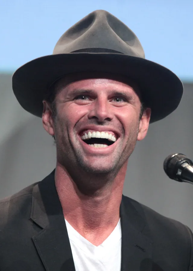 Close photograph of a man laughing broadly, wearing a grey fedora and a dark jacket over a white t-shirt, with a microphone at the edge of the frame.